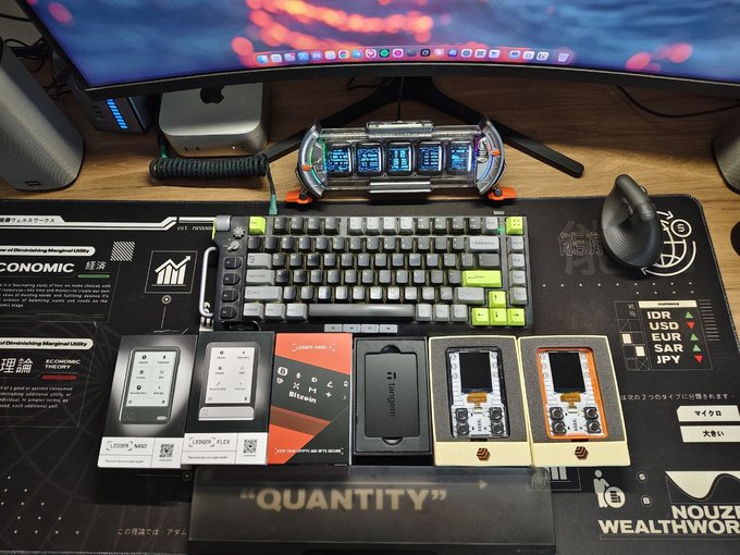 A desk setup featuring a keyboard, mouse, and monitor with several hardware cryptocurrency wallets displayed in their packaging, including the Ledger Nano S Plus, Trezor Safe 3, and what appears to be a "Bitcoin Only" device. The image illustrates the author's concern, showing multiple small crypto hardware wallets alongside a keyboard, suggesting the discomfort of managing valuable collections on such limited-screen devices.