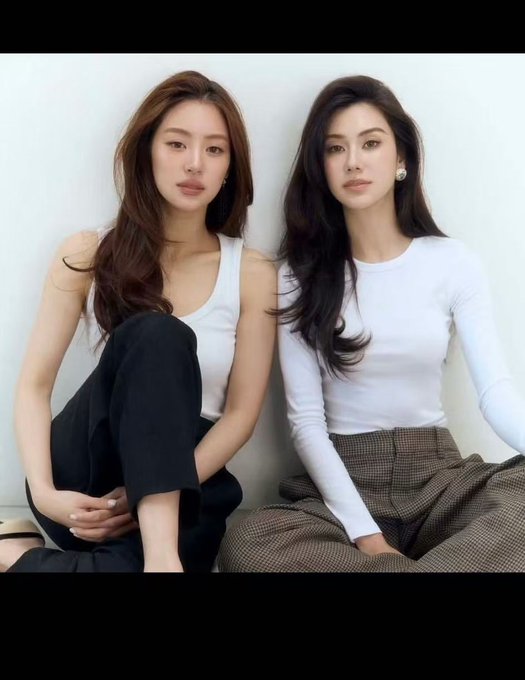 Two women sit side by side against a white background, both with long dark hair; one wears a white tank top and black pants, while the other wears a white long-sleeve top and brown plaid pants.