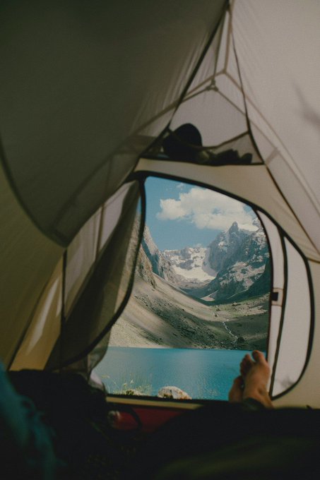 A scenic view from inside a tent looking out at snow-capped mountains and a turquoise alpine lake, with a person's bare foot visible in the foreground.