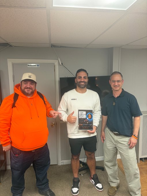 Three men pose for a photo in what appears to be an office setting, with the man in the center holding an award or book titled 'BITCOIN SLASH'.