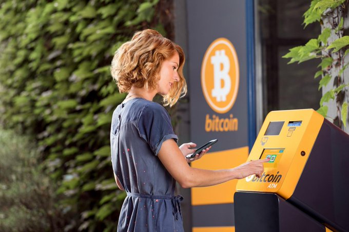 A woman stands next to a Bitcoin ATM, looking down at her phone while interacting with the machine's interface.