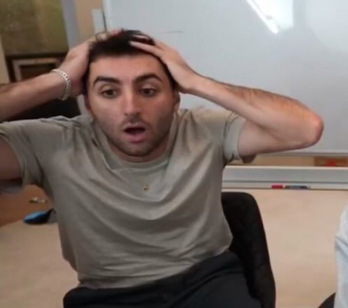 A man in a tan t-shirt sits in a classroom setting, holding his head in both hands with a shocked, dismayed expression.