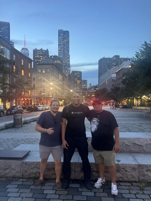 Three men posing together in an urban plaza at dusk with illuminated city buildings and storefronts in the background.