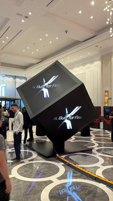 The image features a large, black cube-shaped display with two screens inside of it, one of which has the word "XRP" written on it. The display is situated within an indoor setting, possibly at an event or exhibition. There are several people gathered around the display, some standing and others sitting in chairs nearby. A man can be seen wearing a white shirt among the crowd.