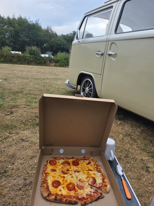 A pepperoni pizza in an open cardboard box sits on the ground in a grassy field, with a white knife and cup nearby, and a vintage cream-colored van parked in the background.
