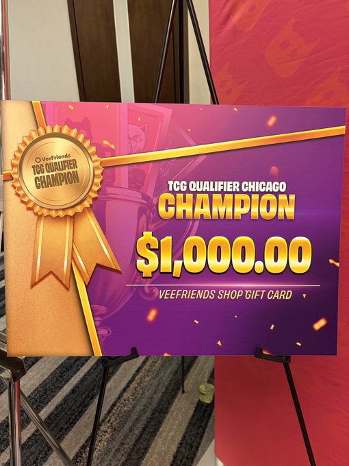 A celebratory poster/sign on an easel announcing a 'TCG QUALIFIER CHICAGO CHAMPION' awarding a $1,000.00 VEEFRIENDS SHOP GIFT CARD, decorated with a gold ribbon medal graphic.