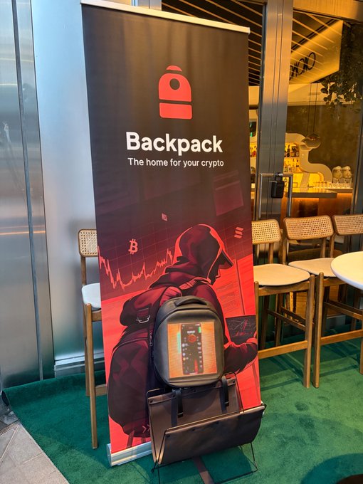 A Backpack crypto company banner reading 'The home for your crypto' displayed indoors with a backpack featuring an electronic device in the foreground.