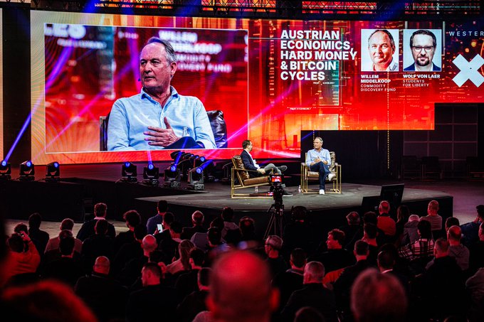 A conference stage featuring two men seated in armchairs with large screens behind them displaying 'AUSTRIAN ECONOMICS, HARD MONEY & BITCOIN CYCLES' with speakers Willem Middelkoop, Founder/Director Discovery Fund, and Yves Vonlaer, Student & FZ Library.