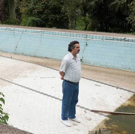 A man in a blue shirt and jeans stands by an empty pool with cracked, blue-tiled walls, hands behind his back.