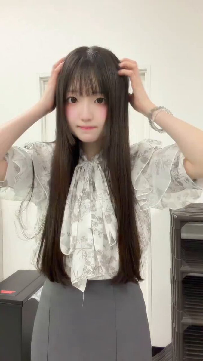 The image features a woman with long brown hair wearing a white blouse, gray skirt, and a bow tie. She has her hands on her head as if she's scratching an itch or adjusting something. The background of the photo shows a door in the room where the woman is standing. There are no other people visible in the image, and it appears to be a simple indoor setting with minimal distractions.