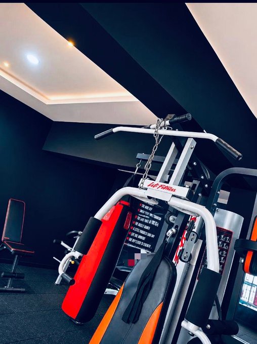 A home or commercial gym interior featuring a multi-station weight machine with red and orange padding labeled 'LIFEFitness', set in a room with black walls and recessed ceiling lighting.