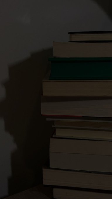 A dimly lit stack of books sits against a dark wall, evoking the quiet focus of returning to study and research after a period of observation. The image complements the post's theme of "locking back in" to dig deeper into selected crypto projects.