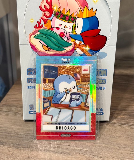 A holographic trading card featuring a penguin character at a sports convention sits in front of a decorative box, labeled 'PUDGY PENGUINS', 'The National Sports Collectors Convention', and 'CHICAGO'.