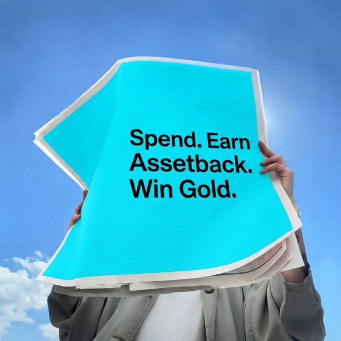 A person holds up a large cyan sheet of paper reading "Spend. Earn Assetback. Win Gold." against a blue sky background, visually reinforcing the post's message about turning everyday payments into rewards and gold through the Assetback program.