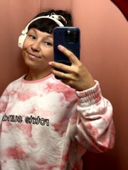 A young person wearing pink headphones and a tie-dye 'Billie Eilish' sweatshirt takes a mirror selfie with a smartphone.