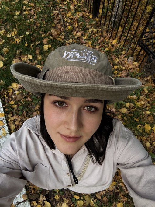 A young woman takes a close-up selfie outdoors while wearing a wide-brimmed olive green hat with 'YORK' embroidered on it and a light-colored jacket.