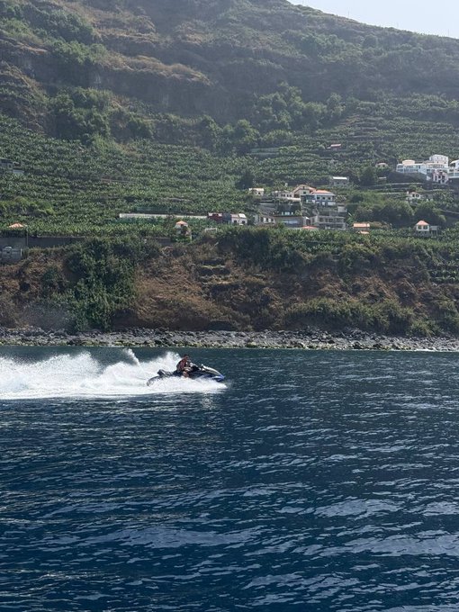 A person rides a jet ski across blue coastal waters with a scenic cliffside village and terraced hillsides in the background, capturing the adventurous "gm to absolutely everyone" energy of starting the day with excitement and good vibes.