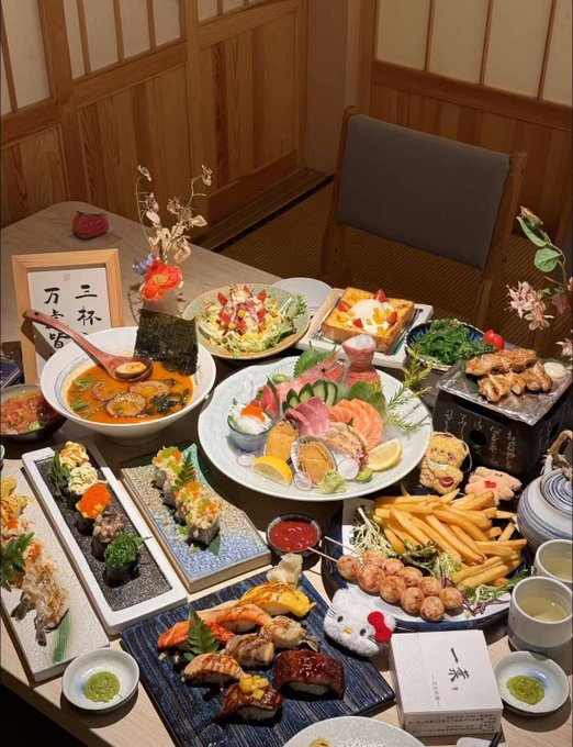 A lavish spread of Japanese cuisine on a wooden table, featuring sushi, sashimi, ramen, tempura, fries, and various other dishes, with a small Hello Kitty figure and a sign with Japanese characters.
