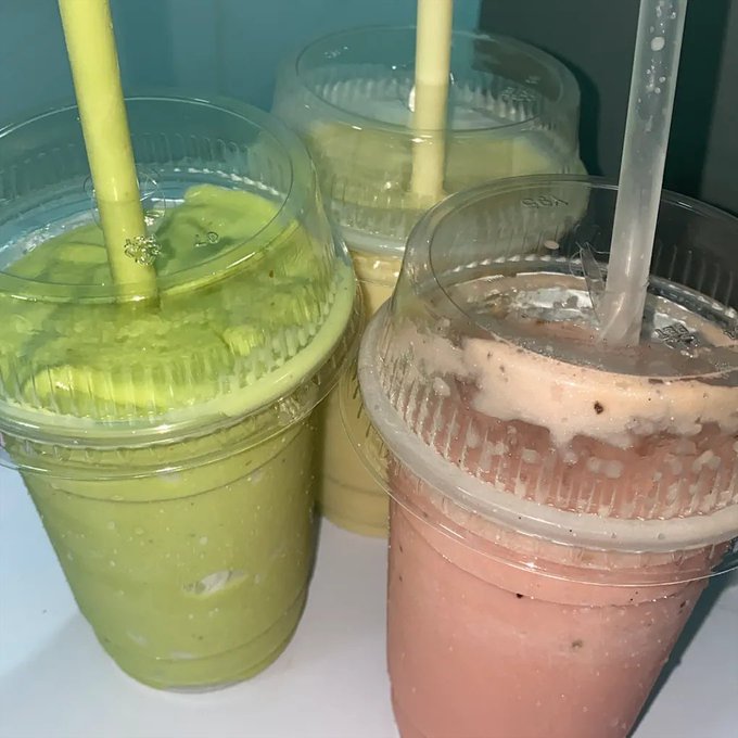 Three plastic cups of smoothies or juices with yellow straws—one green, one yellow, and one pink/brown—sitting on a white surface.