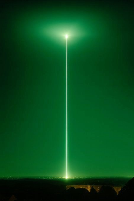 A bright green vertical beam of light shoots up into a green-tinted night sky from a city below.