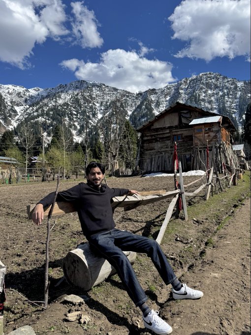 A man sits on a wooden bench in a rural setting, with a snow-capped mountain range and an old wooden cabin in the background.