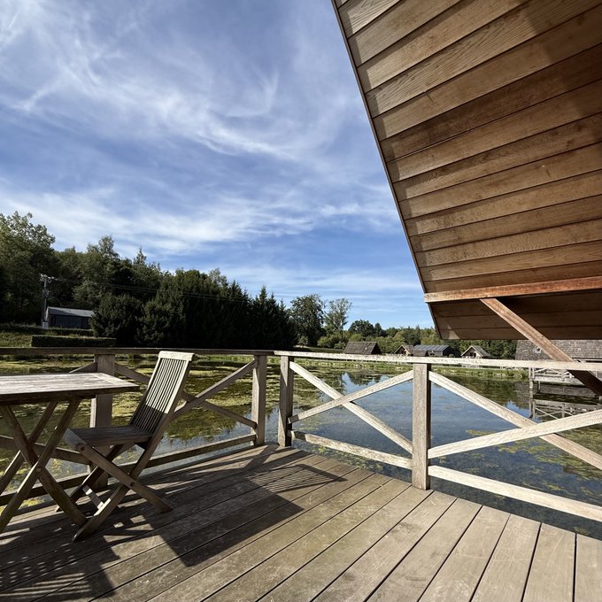 A wooden deck with a chair and railing overlooks a calm pond reflecting the blue sky and trees, with the wooden side of a building visible on the right.