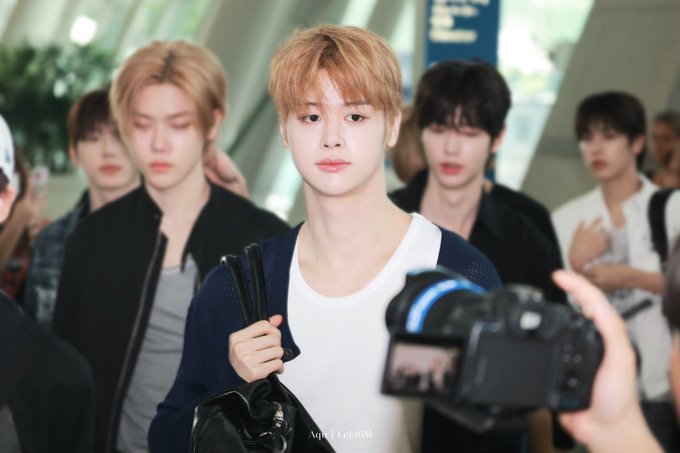 A candid photo of a young man with orange hair wearing a white t-shirt and dark cardigan at an airport, surrounded by other people and a photographer holding a camera.