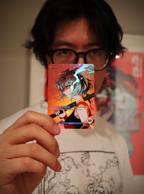 A person with glasses holding up a holographic anime-style trading card featuring a character named 'Shao' with a value of 20.