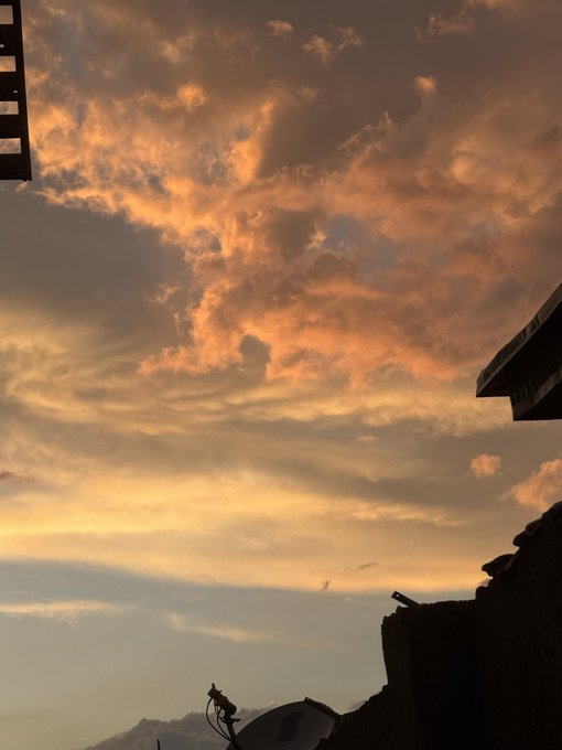 A dramatic sunset sky with orange and peach-colored clouds fills the frame, with the dark silhouettes of rooftops and a satellite dish visible in the lower right corner.