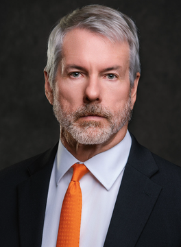 A professional headshot of a middle-aged man with gray hair and a beard, wearing a black suit, white shirt, and bright orange tie, believed to be Michael Saylor, the Strategy (formerly MicroStrategy) executive chairman known for his substantial Bitcoin acquisitions. The bold orange tie visually echoes the bullish sentiment of his continued $BTC accumulation, as referenced in the accompanying post about Strategy's latest $80M Bitcoin purchase.