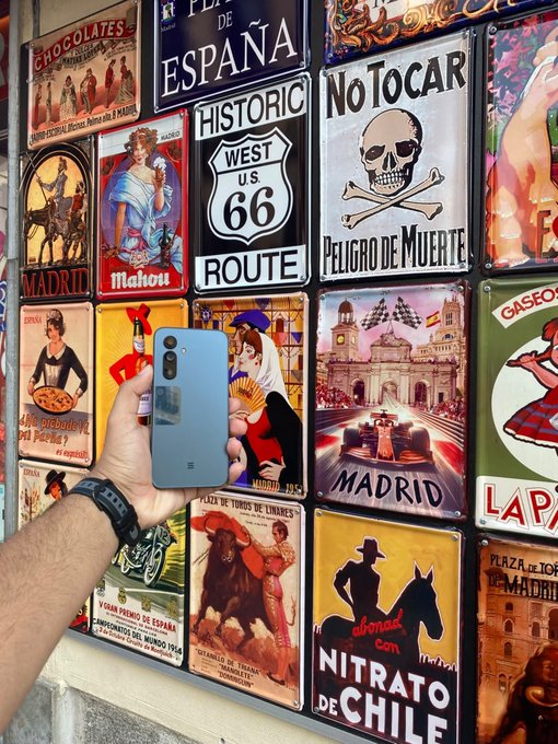 A hand holding a light blue smartphone photographs a colorful display of vintage-style Spanish tin signs featuring designs for 'Plaza de España', 'Historic Route 66', 'No Tocar', 'Madrid', and 'Nitrato de Chile'.