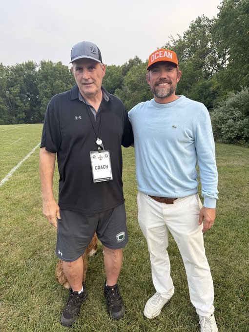 Two men posing on a grassy field; one wears a black Under Armour polo and 'COACH' lanyard, the other wears a light blue sweater and orange 'OCEAN' cap.