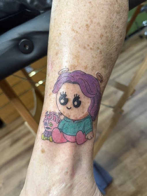 A tattoo on an arm depicting a cartoon-style angel character with purple hair and a halo, sitting next to a small plush toy.