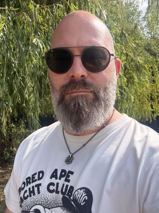 A bald man with a gray beard wearing sunglasses and a white 'Bored Ape Yacht Club' t-shirt poses outdoors in front of a willow tree.