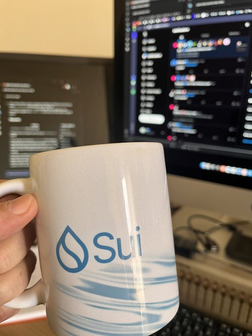 A hand holding a white ceramic mug featuring the blue Sui (SUI) cryptocurrency logo, with computer monitors displaying code in the background.
