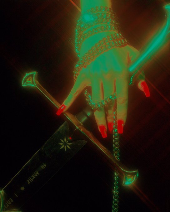 The image features a hand with red nail polish, holding a sword or knife that has a green gemstone on top of it. The background appears to be black, which contrasts with the vibrant colors of the hand's nails and the weapon. The image also contains text that reads "gm folks! wanna thank @OGDSZN for collecting an edition of 'Man Up', a piece about empowerment and cutting thru the BS".