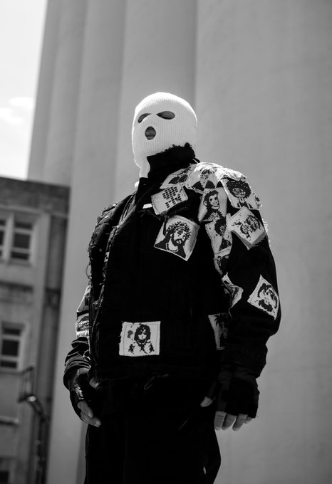 A black-and-white photograph of a person standing against a concrete wall, wearing a white balaclava with eye and mouth holes, and a black bomber jacket adorned with printed patches featuring religious imagery.
