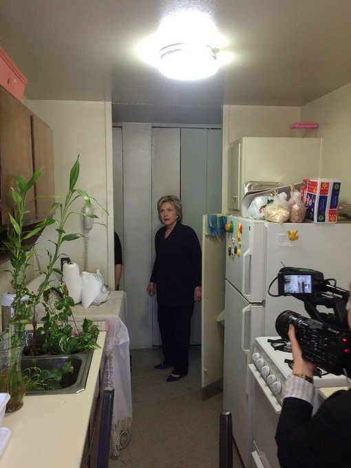 A woman in dark clothing stands in a small kitchen between a refrigerator and cabinets, while a videographer in the foreground films her with a professional camera.