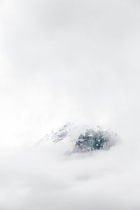 A misty, atmospheric landscape featuring a snow-capped mountain peak emerging from dense clouds and fog, with a desaturated, monochromatic color palette.