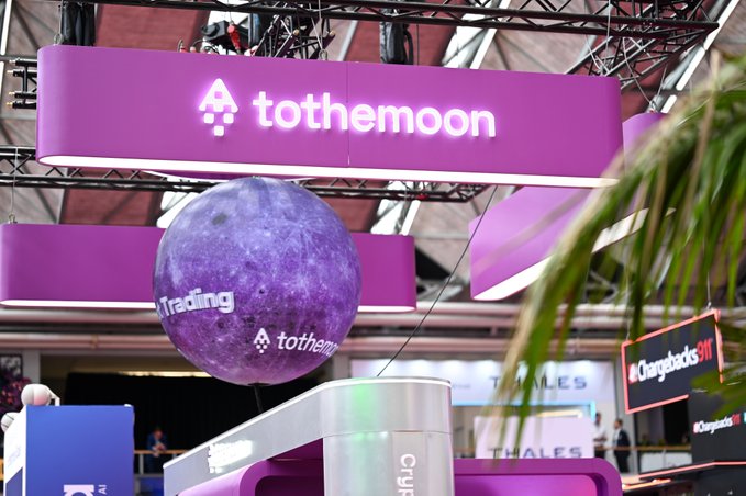 A vibrant purple moon branded with "Trading" and the "tothemoon" logo hangs beneath a matching sign at what appears to be a crypto or fintech conference booth. The display aligns with the post's theme of seamless crypto transactions and scaling opportunities, with a "Chargebacks911" booth visible in the background.