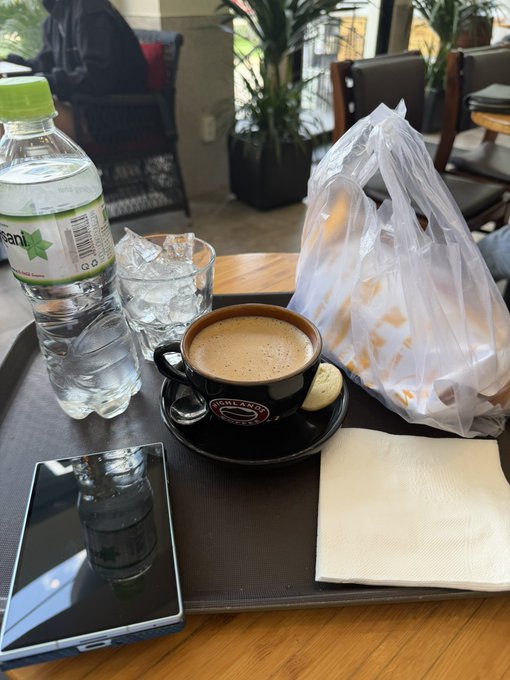 A tray holds a cup of coffee, a bottle of Volvic water, a glass of ice water, a wrapped pastry, and a smartphone resting on a napkin at a cafe.