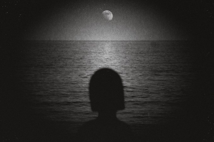 A silhouette of a person looking out over a calm ocean at night with a moon shining in the sky, rendered in black and white.