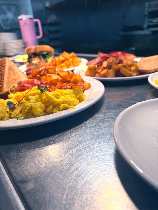 A close-up of a kitchen counter displays plates of food including scrambled eggs with bacon, a sandwich, and home fries on a stainless steel surface.