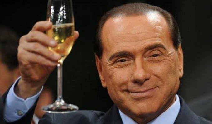 A close-up photo of Italian former Prime Minister Silvio Berlusconi smiling while raising a glass of white wine.
