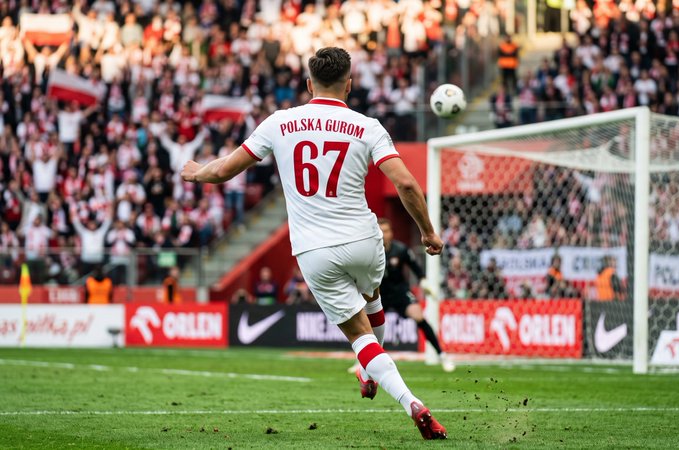 A soccer/football image showing a player in a white jersey with 'POLSKA GURUM 67' running on a green field during a match, with a packed stadium and Orlen advertising boards in the background.