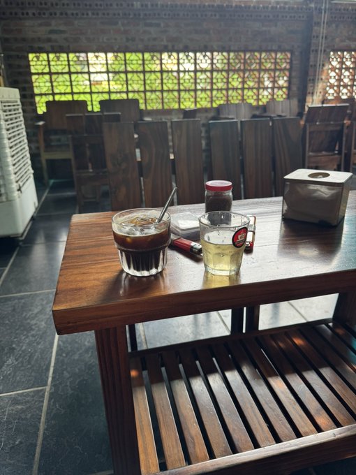 A wooden table in a cafe holds a glass of iced dark coffee, a glass of light-colored beer with a red logo, a jar of dark preserve, a lighter, and a tissue box, with empty wooden chairs visible in the background.