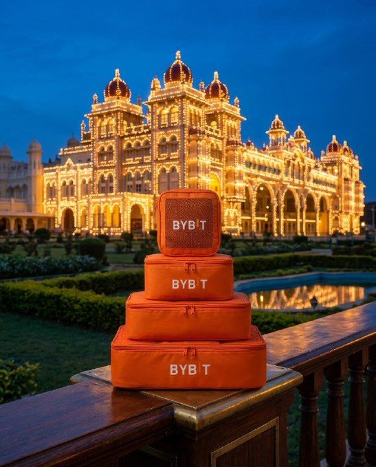 A travel/luggage promotional image showing stacked orange 'BYB'T branded suitcases and a matching speaker on a wooden railing, with the illuminated Mysore Palace in India lit up in golden lights at dusk in the background.
