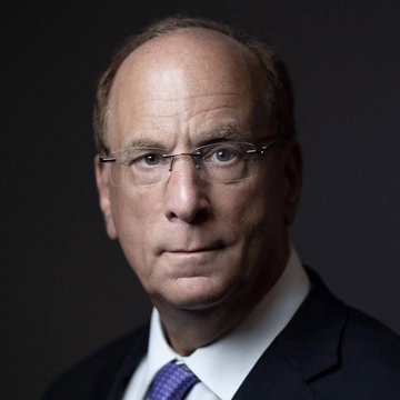 The image shows an elderly man wearing glasses sitting against a dark background. The close-up of his face, which makes up most of the photo, gives a clear view of his features. The photograph has a professional, authoritative look to it and is presented in black and white.