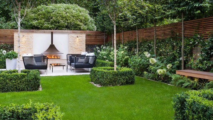 A landscaped garden with a lush green lawn, featuring an outdoor seating area with a fireplace and black wicker chairs, surrounded by hydrangeas, hedges, and wooden fencing.