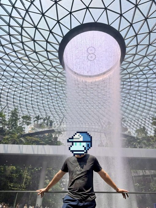 A man wearing a black t-shirt stands in front of the iconic Rain Vortex waterfall inside Jewel Changi Airport's glass dome, with his face obscured by a pixelated graphic.